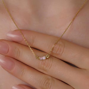 May include: A gold necklace with a small, rectangular pink gemstone pendant. The necklace is shown on a person's hand, highlighting the fine chain and the pendant's subtle sparkle. The jewelry is a simple and elegant accessory.