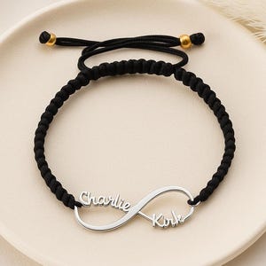May include: A black braided cord bracelet with a silver infinity symbol. The infinity symbol has the names "Charlie" and "Kirk" engraved on it. The bracelet has gold-colored beads at the ends.