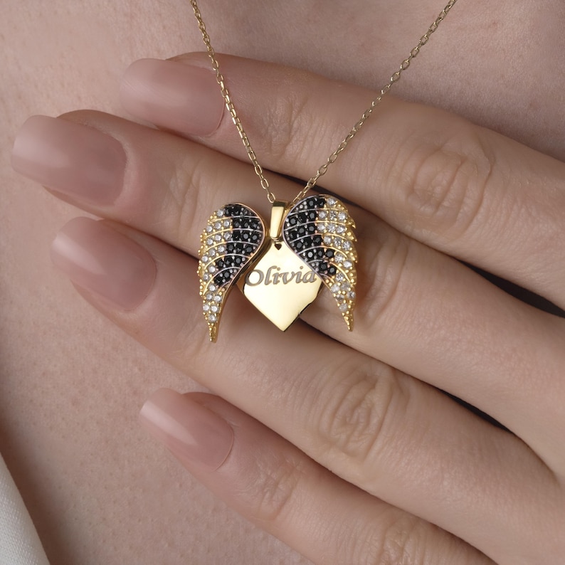 May include: A gold-colored necklace with a pendant shaped like angel wings. The wings are detailed with black and clear stones. A heart-shaped tag in the center is engraved with the name "Olivia". The necklace is displayed against a neutral background.