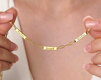 May include: A gold-colored necklace with three rectangular nameplates. The names "Audrey", "Joseph", and "Evelyn" are engraved on the plates. The necklace is held by a person, and the background is a soft, neutral color.