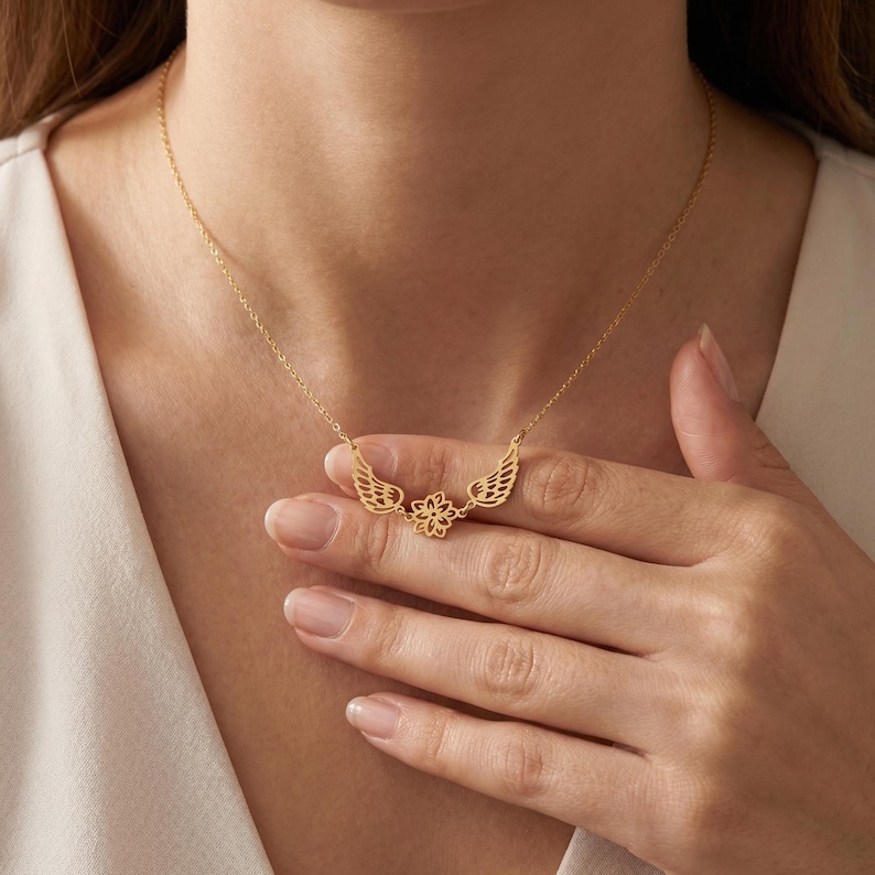 May include: A gold-colored necklace featuring a delicate design of wings and a flower. The necklace is worn around the neck and is held by a hand. The wings are on either side of the flower. The necklace is a fine piece of jewelry.