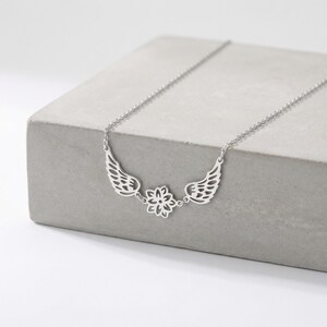 May include: A silver-colored necklace featuring a delicate design of angel wings and a central floral element. The necklace is displayed on a gray, concrete surface, showcasing its intricate details and elegant style. The chain is a standard length.