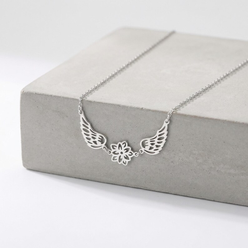 May include: A silver-colored necklace featuring a delicate design of angel wings and a central floral element. The necklace is displayed on a gray, concrete surface, showcasing its intricate details and elegant style. The chain is a standard length.