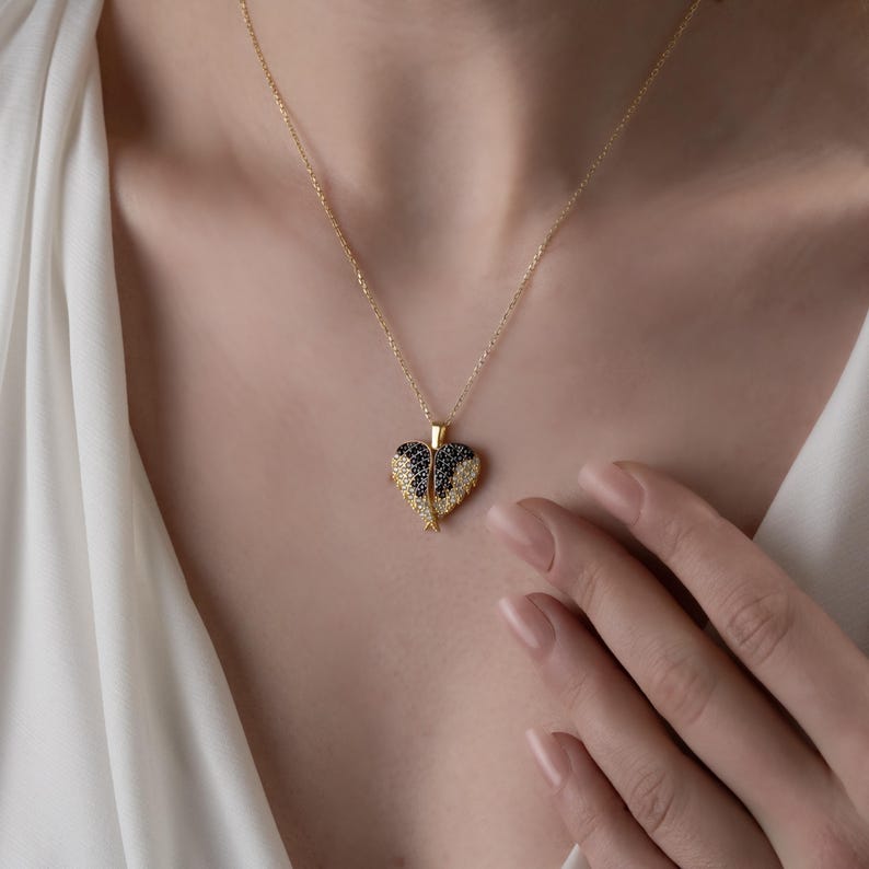 May include: A gold-colored necklace with a heart-shaped pendant. The pendant features a black and white gemstone design. The necklace is worn around the neck.