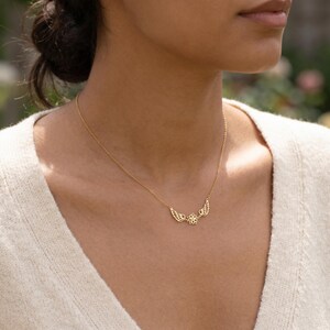 May include: A gold-colored necklace featuring a central flower design flanked by two sets of wings. The necklace is delicate and appears to be made of a thin chain. The necklace is worn on a person.