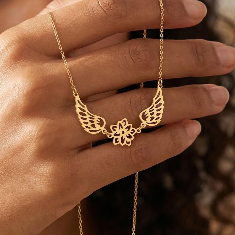 May include: A gold-colored necklace featuring a floral pendant flanked by two openwork wings. The delicate chain and pendant are displayed against a blurred background, highlighting the jewelry's intricate design.