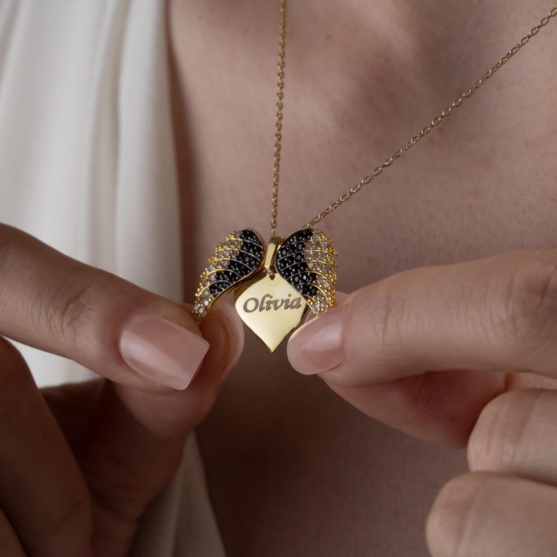 May include: A gold-colored necklace featuring a pendant with the name "Olivia" engraved on a heart-shaped tag. The pendant is flanked by two wing-shaped elements, adorned with black and clear gemstones. The necklace is held by a person.