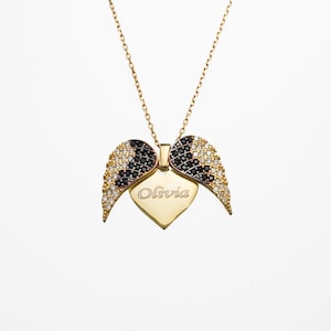 May include: A gold-colored necklace with a heart-shaped pendant and wing accents. The pendant is engraved with the name "Olivia". The wings are adorned with black and white gemstones, and the necklace has a delicate chain.