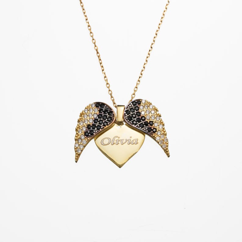 May include: A gold-colored necklace with a heart-shaped pendant and wing accents. The pendant is engraved with the name "Olivia". The wings are adorned with black and white gemstones, and the necklace has a delicate chain.