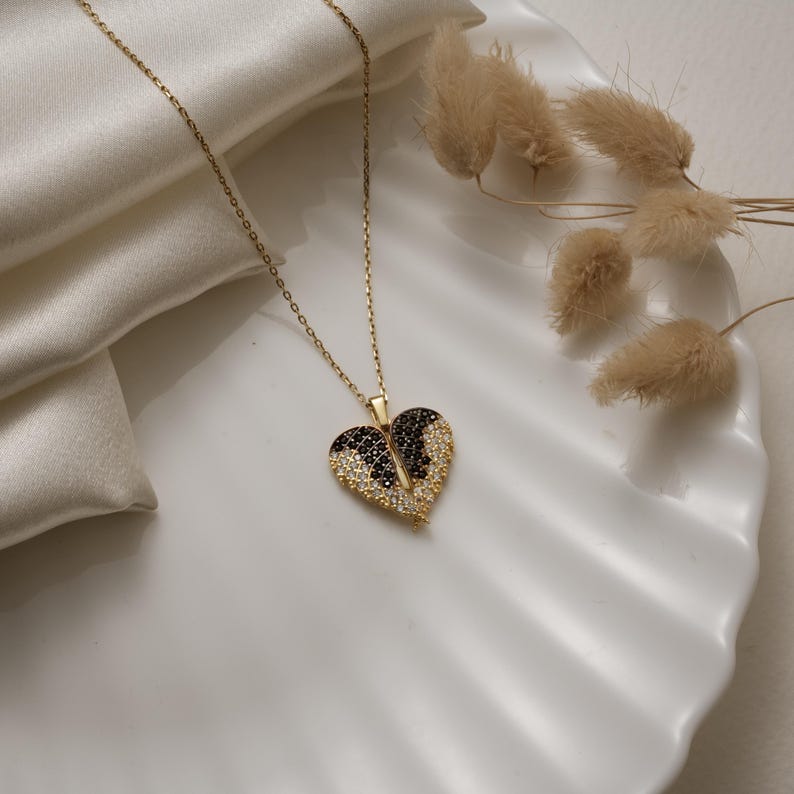 May include: A gold-colored necklace with a heart-shaped pendant. The pendant is split in half, with one side featuring black stones and the other side featuring clear stones. The necklace is displayed on a white surface.