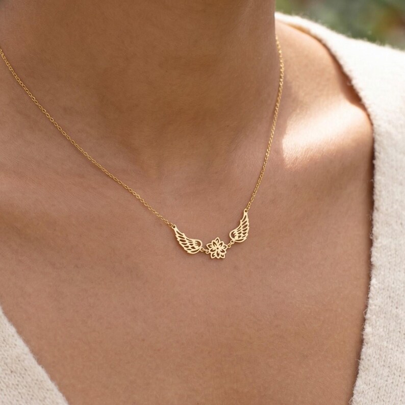May include: A gold-colored necklace featuring a delicate design. The necklace has a chain with a pendant composed of two stylized wings flanking a floral element. The wings and flower are intricately detailed, creating an elegant and refined aesthetic.