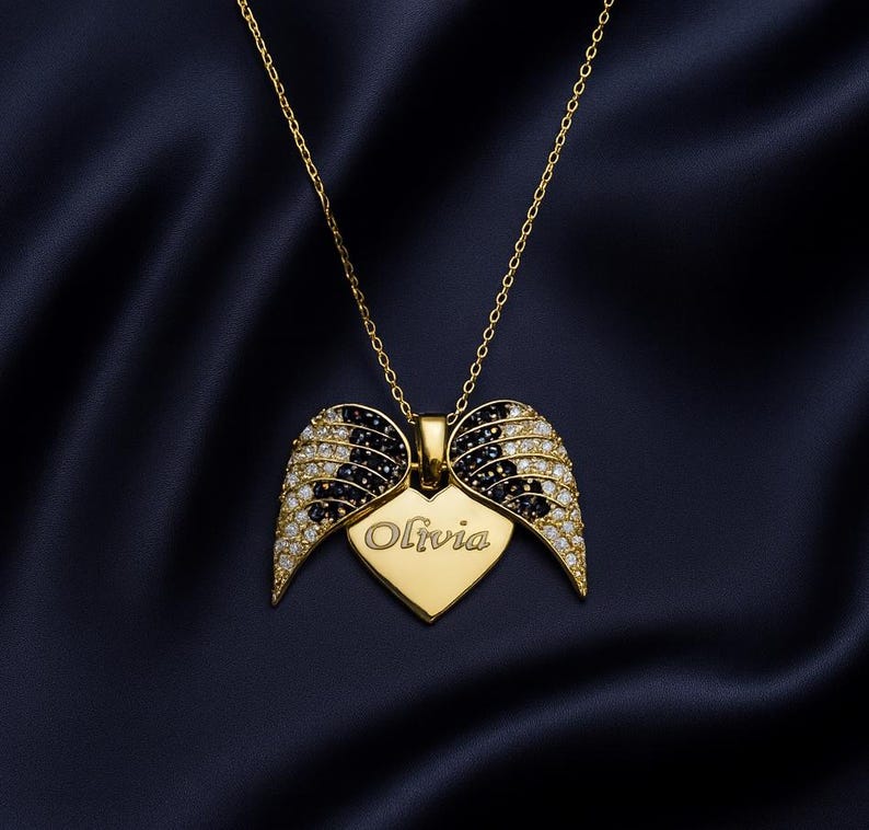 May include: A gold-colored necklace with a heart-shaped pendant and wing design. The pendant has the name "Olivia" engraved on it. The wings are adorned with black and clear gemstones, creating a sparkling effect. The necklace hangs on a dark blue background.