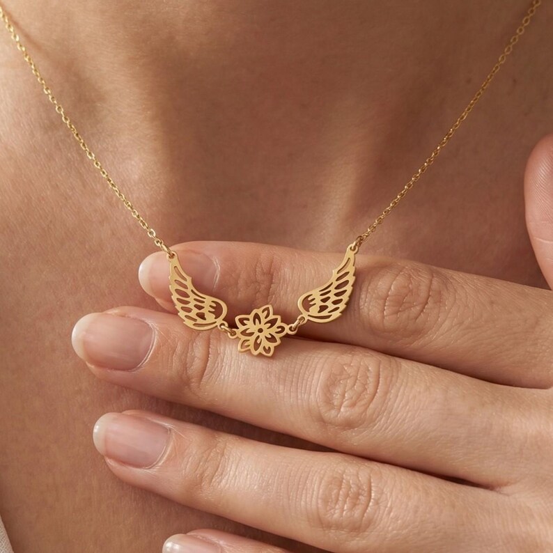 May include: A gold-colored necklace featuring a delicate design. The necklace has a chain with a pendant that includes stylized wings on either side of a floral element. The necklace is shown on a person's neck.
