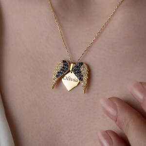 May include: A gold-colored necklace with a pendant shaped like angel wings. The wings are adorned with black and clear gemstones. A heart-shaped tag with the name "Olivia" is suspended from the wings. The necklace is displayed against a skin-toned background.
