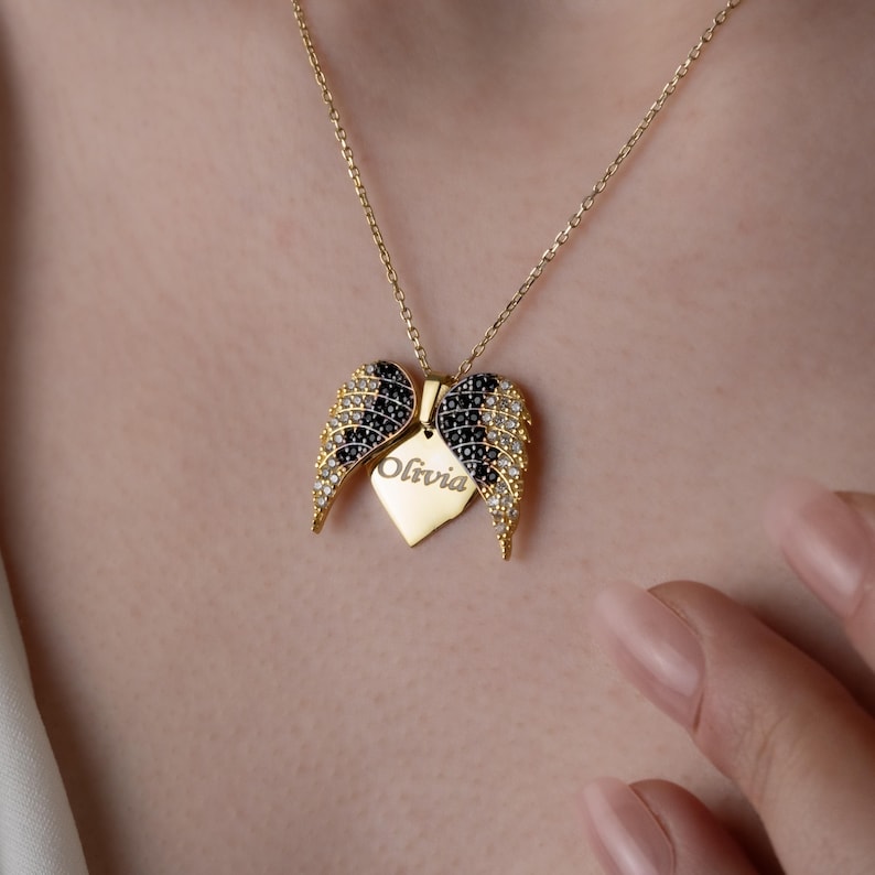 May include: A gold-colored necklace with a pendant shaped like angel wings. The wings are adorned with black and clear gemstones. A heart-shaped tag with the name "Olivia" is suspended from the wings. The necklace is displayed against a skin-toned background.