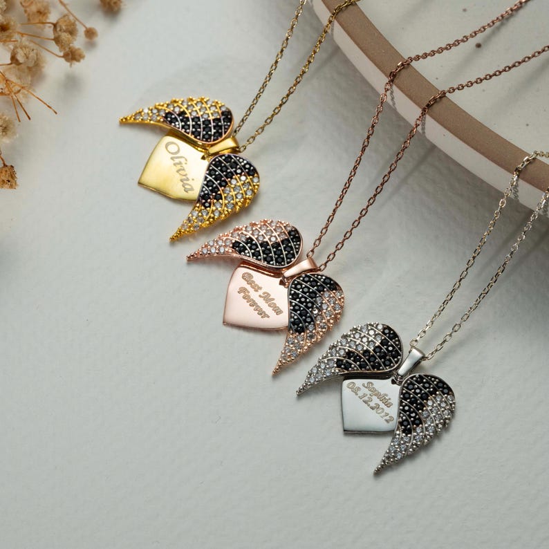May include: Four necklaces with heart-shaped pendants and wing accents. The pendants are gold, rose gold, and silver, with black and clear crystal detailing. Each heart has engraved text, including names and dates.