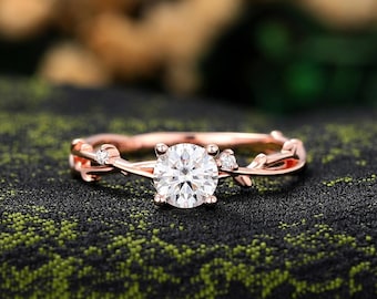Könnte beinhalten: Roségoldener Ring mit einem großen, runden, klaren Mittelstein und kleineren Akzentsteinen. Das Band hat ein zartes Wein- und Blattdesign. Der Ring ist vor einem dunklen, strukturierten Hintergrund platziert.