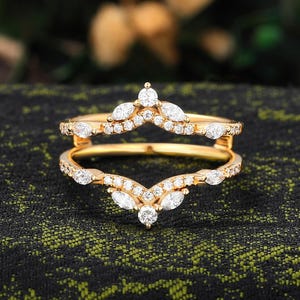 Può includere: Una protezione per anello color oro con un design a forma di V. L'anello è adornato con numerose piccole gemme trasparenti e pietre più grandi con taglio a marquise. L'anello è posto su uno sfondo scuro e strutturato.
