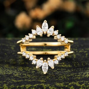 Op de afbeelding: Een goudkleurige ringbeschermer met een dubbelbandontwerp. De ring heeft een reeks kleine, ronde en marquise-geslepen heldere edelstenen die in een decoratief patroon zijn gezet. De ring is ontworpen om met een andere ring te worden gedragen.