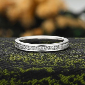 May include: A silver ring featuring a continuous line of small, clear gemstones. The ring is set against a dark green and black textured backdrop, with a blurred background of yellow and green.