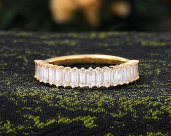 May include: A gold-colored ring with a band of rectangular, clear stones. The ring is set against a dark, textured background, with a blurred, golden-toned backdrop.
