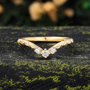 Puede incluir: Un anillo dorado con un diseño curvo, con tres pequeñas piedras preciosas claras. La banda tiene un patrón de hojas. El anillo está sobre un fondo oscuro y texturizado.
