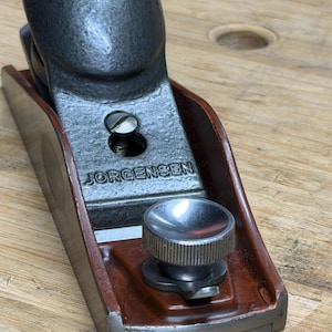 May include: A Jorgensen hand plane, featuring a gray metal body and a dark brown wooden base. The plane has a rounded top and a silver adjustment knob. The word "JORGENSEN" is stamped on the metal.