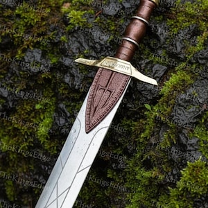 May include: A detailed sword with a silver blade and a brown handle. The hilt features gold accents and the word "ANAKAVDIVINY". The blade has etched geometric patterns. The sword rests on a mossy surface.
