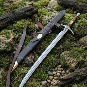 May include: A steel sword with a brown wooden handle and a silver pommel, featuring a crossguard with a bird emblem. The sword is accompanied by a dark brown leather scabbard with brass accents and a long leather strap for carrying the weapon.