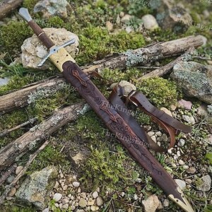 May include: A sword with a brown leather-wrapped handle and a silver-colored crossguard. The sword is housed in a dark brown leather scabbard featuring intricate embossed knotwork patterns, brass-colored metal accents, and a leather strap.
