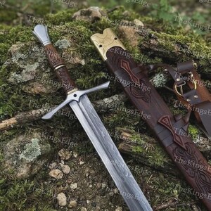 May include: A steel sword with a patterned blade, a brown wooden grip, and a silver crossguard. Beside it lies a matching brown wooden scabbard featuring intricate carvings, a brass-colored metal throat, and leather straps with metal rings.