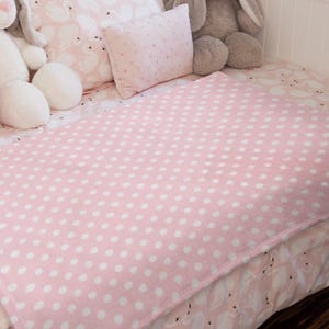 May include: A pink blanket with white polka dots rests on a bed with swan-patterned bedding. Two plush bunny toys and a small floral-patterned pillow are also present, creating a soft nursery setting.