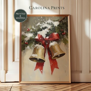 Pode incluir: Uma impressão de arte emoldurada intitulada CAROLINA PRINTS apresentando uma pintura de dois sinos dourados pendurados em um galho de pinheiro coberto de neve com bagas vermelhas e um grande laço vermelho em um fundo creme claro.