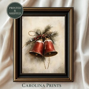 Può includere: Una stampa artistica incorniciata raffigurante due campane rosse con bordi dorati, spolverate di neve bianca, legate insieme con spago e rami di pino con bacche rosse. La cornice nera ha un bordo dorato. Testo: Printed & Shipped e Carolina Prints.