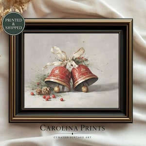 Può includere: Una stampa artistica incorniciata raffigurante due campane natalizie rosse legate da un nastro color crema, arricchite da aghi di pino, piccole pigne e bacche rosse. L'opera è racchiusa in una cornice decorativa di colore nero e oro.