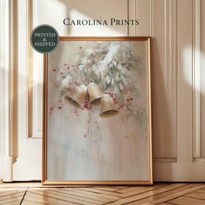 Pode incluir: Uma impressão de arte emoldurada intitulada Carolina Prints apresentando uma ilustração suave e pictórica de três sinos dourados pendurados em ramos de pinheiro com bagas vermelhas. A obra tem um fundo claro e suave com pinceladas visíveis.