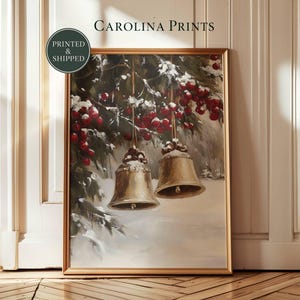 Könnte beinhalten: Ein gerahmter Kunstdruck mit dem Titel CAROLINA PRINTS zeigt ein Gemälde von zwei goldenen Glocken, die an einem mit Schnee und roten Beeren bedeckten Tannenzweig hängen. Die Szene zeigt eine Winterlandschaft mit weißem Boden und grünem Laub.