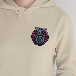 May include: Beige hoodie with a front pocket and drawstring hood. The hoodie features a colorful embroidered owl design on the upper left chest. The owl design incorporates shades of blue, pink, and purple, creating a vibrant contrast against the neutral background.