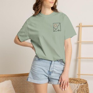 May include: A light green t-shirt with a small square graphic on the upper left chest. The graphic features a red circle and black lines. The shirt is paired with light blue denim shorts.