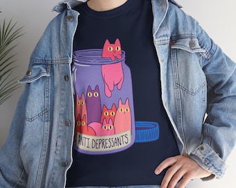 May include: Navy blue t-shirt featuring a whimsical illustration of cartoon cats inside a purple jar labeled "ANTI DEPRESSANTS". The cats are in shades of pink and purple. A blue lid is next to the jar. The shirt is a casual wear item.