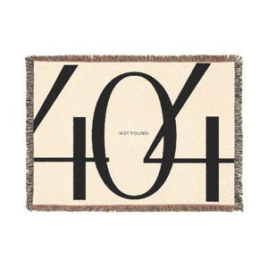 May include: Cream-coloured woven throw blanket featuring a large black "404" design and the words "NOT FOUND" in the centre. The blanket has a fringed edge in shades of brown and red.