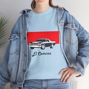 May include: Pale blue t-shirt with a black and white graphic of an El Camino car, set against a red stripe. The words "El Camino" are written in a cursive font below the car. The t-shirt is worn with a denim jacket.