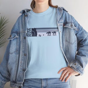 May include: Pale blue t-shirt with a parody design. The design includes the text "THE NORTH ASE" with a mountain graphic, and AT-AT walker silhouettes. The shirt is worn with a denim jacket.