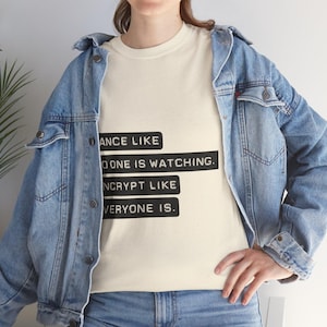 Op de afbeelding: Crèmekleurig T-shirt met de tekst "DANCE LIKE NO ONE IS WATCHING. ENCRYPT LIKE EVERYONE IS." Het shirt wordt gedragen met een lichtblauwe spijkerjas en jeans. De tekst is in een vetgedrukte, schreefloze lettertype.