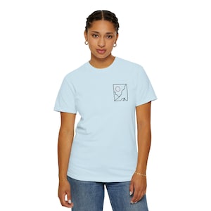 Peut inclure: Un t-shirt bleu clair avec un petit dessin au trait noir d'un soleil, de montagnes et d'un carré. Le t-shirt est en matière douce et est porté.