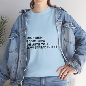 May include: Light blue t-shirt with the text "YOU THINK I'M COOL NOW WAIT UNTIL YOU SEE MY SPREADSHEETS" in black. The shirt is worn under a light blue denim jacket.
