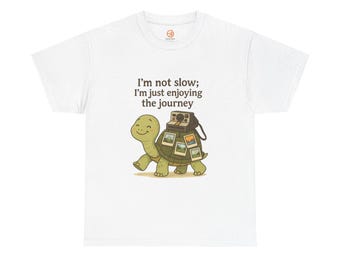 May include: White t-shirt featuring a cartoon turtle carrying a vintage camera and several photos. The text "I'm not slow; I'm just enjoying the journey" is printed above the turtle. The shirt has a crew neck and short sleeves.