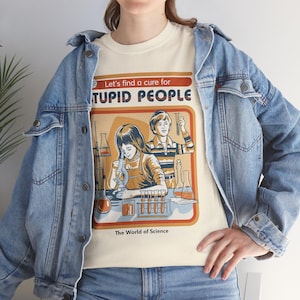May include: Cream-colored t-shirt with a retro graphic featuring two figures in a lab setting, test tubes, and the text "Let's find a cure for STUPID PEOPLE" and "The World of Science". The shirt is worn with a light blue denim jacket.
