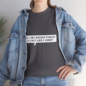 May include: A dark gray t-shirt with the text "DO I GET BONUS POINTS IF I ACT LIKE I CARE?" in a white pixelated speech bubble. The person is wearing a blue denim jacket.