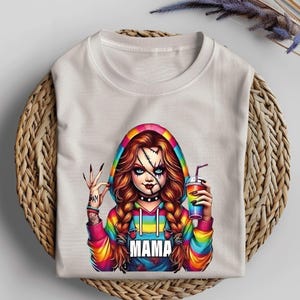 May include: White t-shirt featuring a colorful graphic of a doll-like character with stitched facial details, holding a drink. The character wears a rainbow hoodie with the word "MAMA" on the front. The character has long, braided hair and a tattoo on the arm.